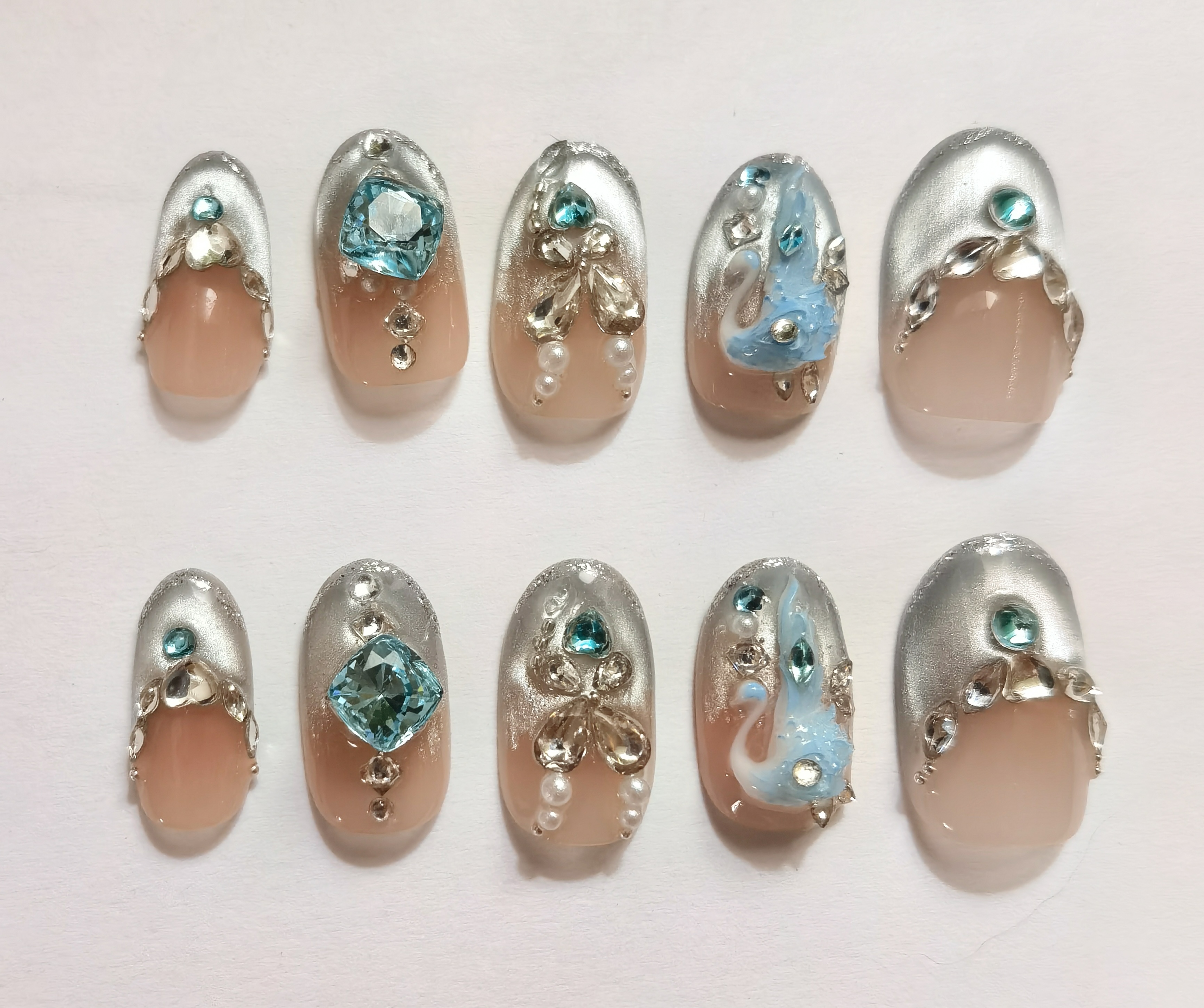 24-piece full set of luxurious almond-shaped press-on nails featuring intricate 3D swan art, sparkling blue and clear rhinestones, and delicate pearl embellishments on a nude and shimmery silver base.