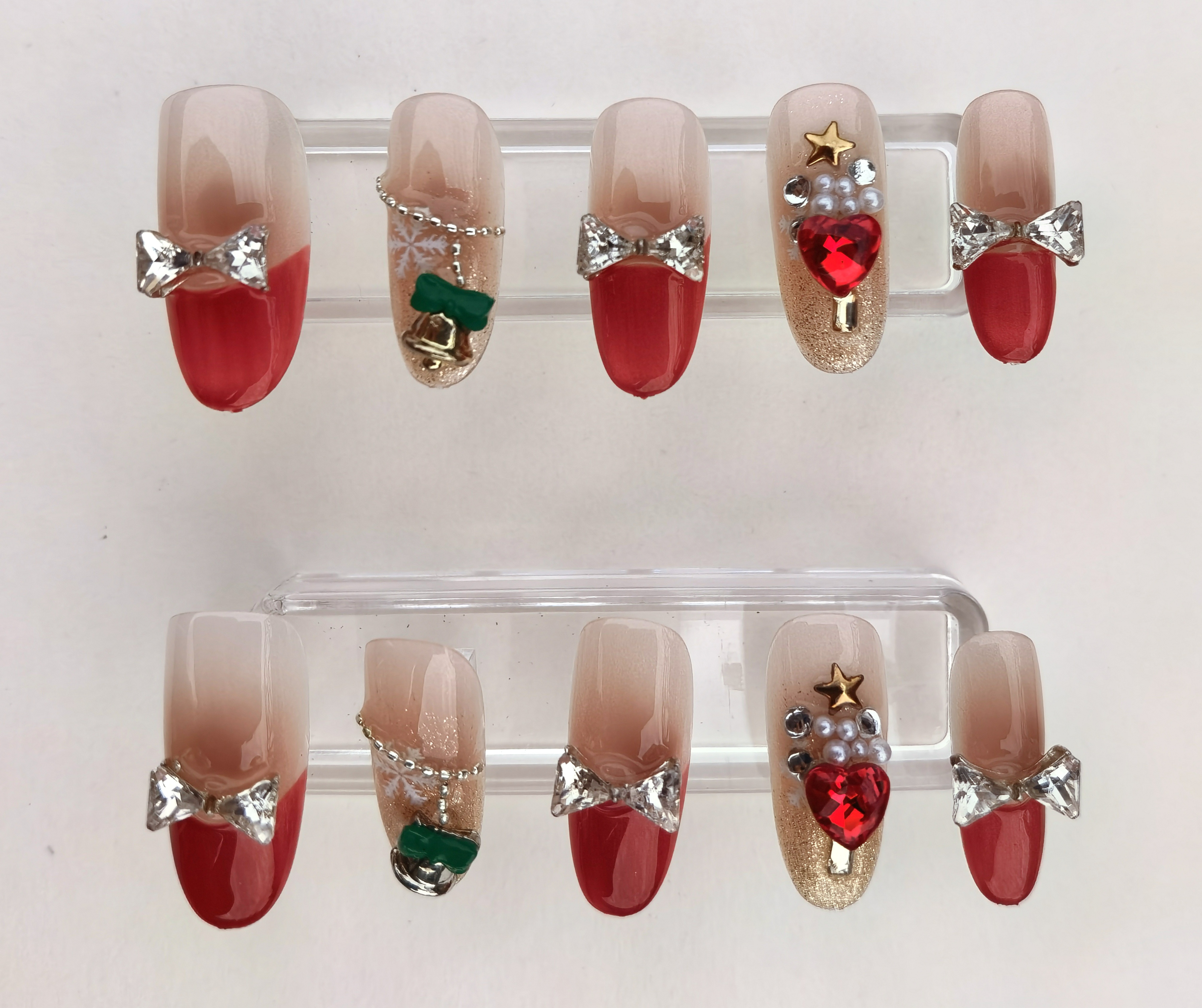 A 24-piece full set of Tenth Touch press-on nails featuring a festive holiday design with glossy red tips, glitter accents, and intricate 3D embellishments including rhinestone bows, hearts, stars, pearls, snowflakes, and bells.
