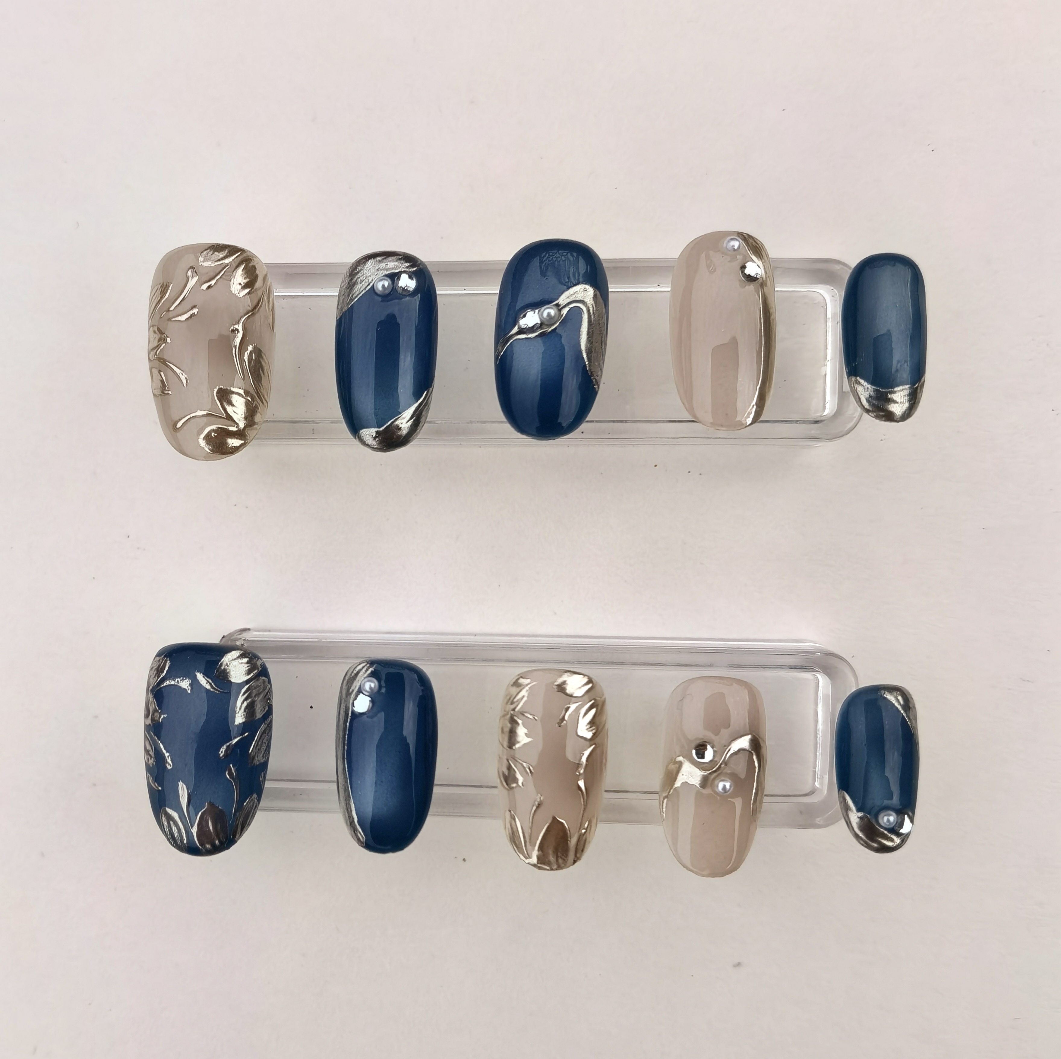 A 24-piece full set of medium oval press-on nails featuring glossy deep blue and nude bases with intricate silver 3D floral and abstract designs, accented with delicate pearls.